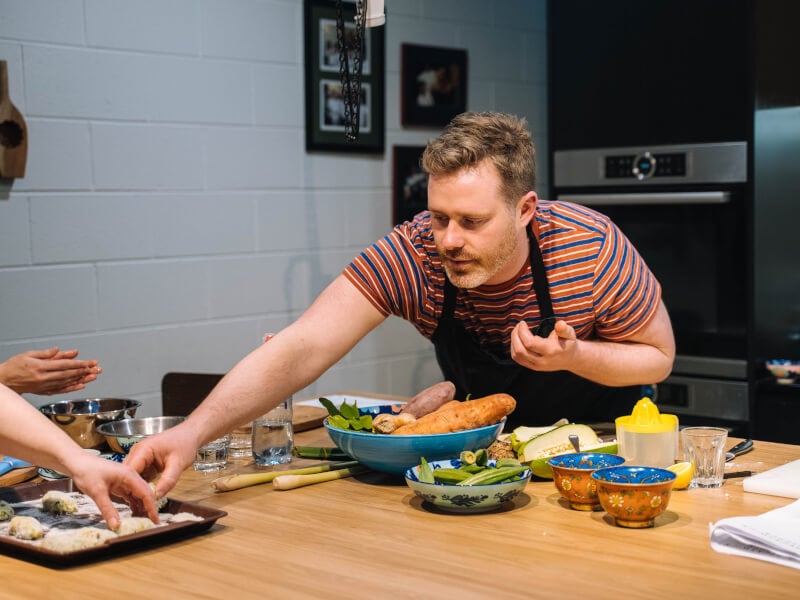 A man stretching across a table to reach some ingredients at a cooking class.